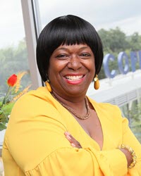 Brown skin woman with black-haired bob is smiling in front of a window while wearing a yellow v-neck blouse, gold necklace, and dangly yellow earrings