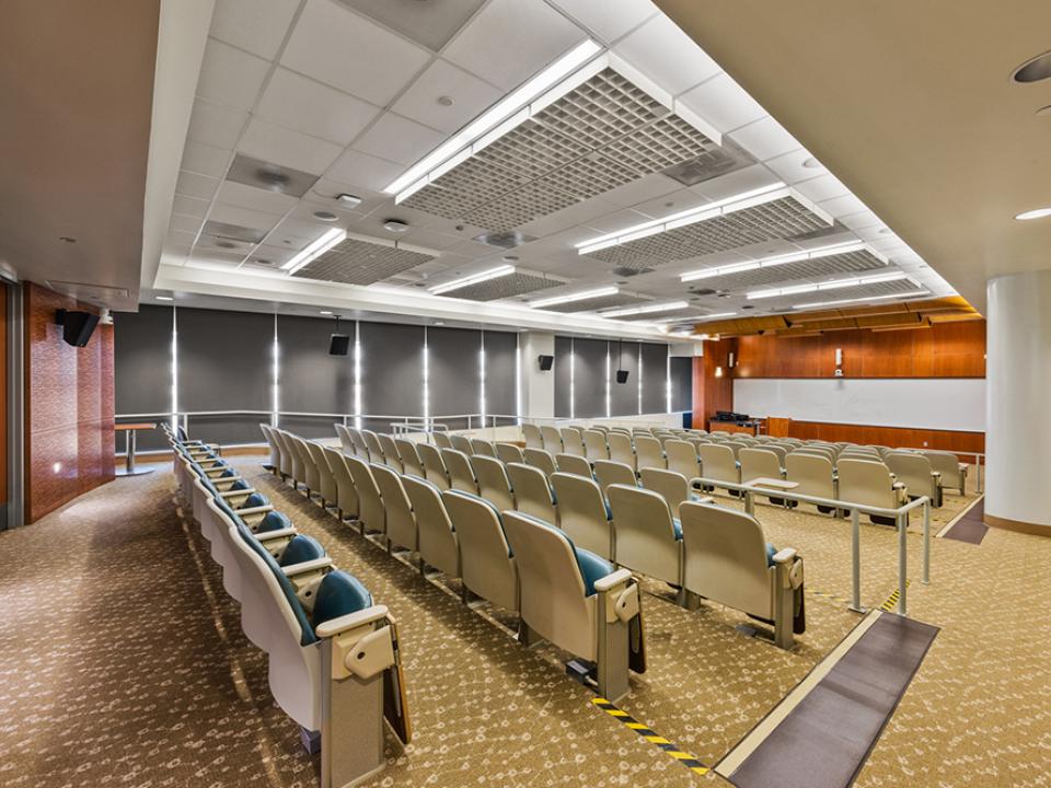 Room 120 lecture room with projector and full room-width screen. Tiered seats are cushioned and include power outlets