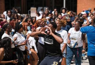 A student wearing a black Coppin State t-shirt cups his hands around his mouth and shouts enthusiastically while surrounded by a large, energetic crowd of students during an outdoor campus event.